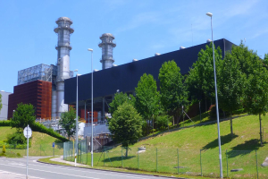 Großes Gebäude mit zahlreichen Fenstern, als Kraftwerk durch Beschilderung identifiziert, umgeben von Straßeninfrastruktur, Fahrzeugen, Grünflächen und einem bewölkten Himmel.