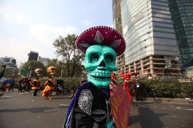 Ein Tag der Toten Umzug in Mexiko-Stadt mit Menschen in Kostümen, die Gegenstände halten und einen klaren blauen Himmel im Hintergrund.
