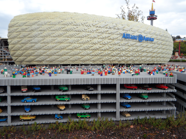 Ein großes Lego-Modell des Allianzstadions in München, Deutschland, mit einer Gruppe von Menschen davor, umgeben von Bäumen, Pfählen, einem Turm und Pflanzen unter einem bewölkten Himmel.