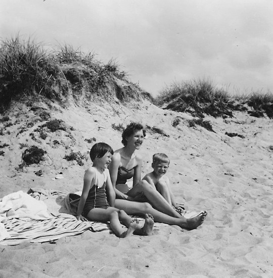 Schwarz-weiß-Foto einer Frau in einem Bikini und zwei Kindern in Badebekleidung, die auf einer Decke am Strand sitzen, mit Sand, Gras und Himmel im Hintergrund.