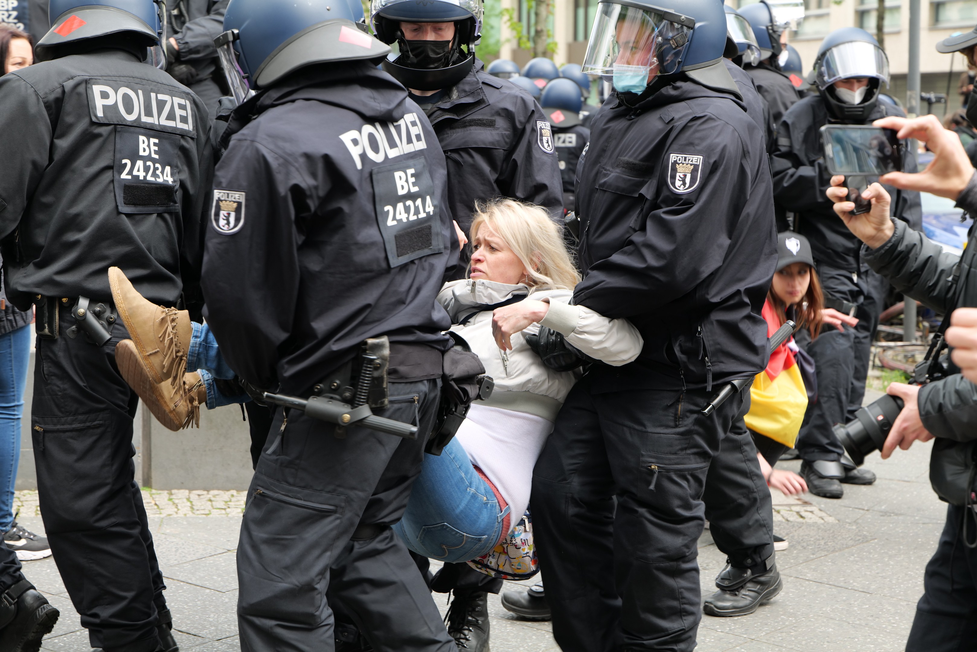 Eine Gruppe von Polizisten in Schutzausrüstung eskortiert eine Frau in einem weißen und blauen Kleid, wobei ein Polizist ihren Arm hält, während ein Fotograf die Szene rechts davon aufnimmt, vor einer Kulisse aus Gebäuden, Bäumen und Himmel.