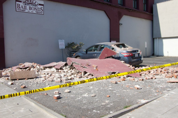 Eine zerbrochene Ziegelwand liegt auf dem Boden mit verstreuten roten Ziegeln, einem geparkten Auto, einer Topfpflanze und einem Gebäude mit einer "Kautionsbonds"-Aufschrift.