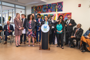 Eine Gruppe von Menschen, die vor einem Rednerpult mit Mikrofon und einer Tafel mit der Aufschrift "Sancho-Stadtrat tritt dem Stadtrat bei" steht und sitzt, mit Flaggen, Feuerlöscher und Deckenleuchten an der Rückwand.