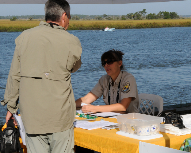 Ein Beamter sitzt an einem Tisch mit Papieren vor einem See, w├Ąhrend ein Mann vor einer Gruppe von Menschen steht.