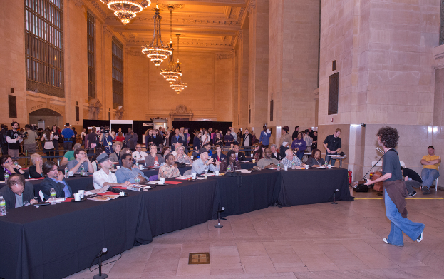 Ein Konzertbild mit einer Menge, einem Mann, der auf der rechten Seite Gitarre spielt und singt, einer Wand und Fenstern im Hintergrund, Lichtern oben und einem Boden unten.