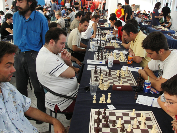 Eine Gruppe von Personen spielt Schach mit einem Mann in einem blauen Hemd auf der linken Seite, ein Fenster in der oberen rechten Ecke und ein Tisch mit einem Glas, einem Schachbrett, Schachfiguren, einer Coke, Papier, einem Stift und einer Tischdecke.