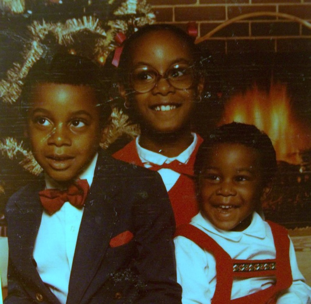 Drei Kinder lachen vor einem Weihnachtsbaum mit einem Kamin aus Feuerholz auf der rechten Seite.