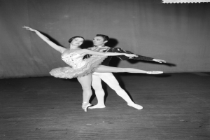 Ein Schwarz-Weiß-Foto eines Mannes in einem weißen Hemd und schwarzen Hosen und einer Frau in einem unbekannten Outfit, die anmutig und perfekt synchron auf einer Bühne tanzen, mit einem Vorhang im Hintergrund.