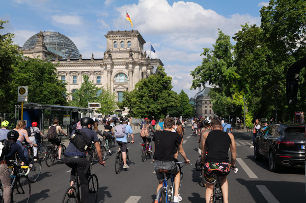 Eine Gruppe von Menschen, die auf Fahrrädern eine von Bäumen gesäumte Straße in Berlin, Deutschland, entlangfahren, mit Gebäuden auf beiden Seiten, einer Bushaltestelle auf der rechten Seite und einer Fahne, die auf einem der Gebäude weht, unter einem bewölkten Himmel.