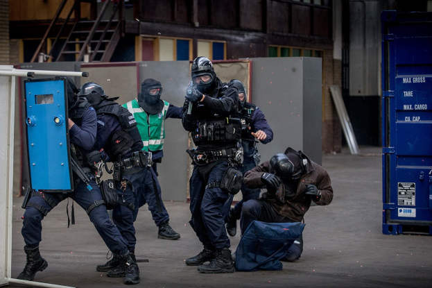 Polizeibeamte in Einsatzausrüstung stehen in einer Reihe vor einem Gebäude, einige halten Waffen, mit einer Person im Vordergrund und Metallobjekten, Treppen und einer Wand im Hintergrund.