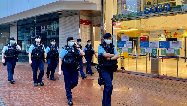 Eine Gruppe von Polizisten in Masken geht eine Straße entlang vor einem Glasgebäude, wobei auf der rechten Seite ein Geschäft mit Plakaten an den Glaswänden zu sehen ist.