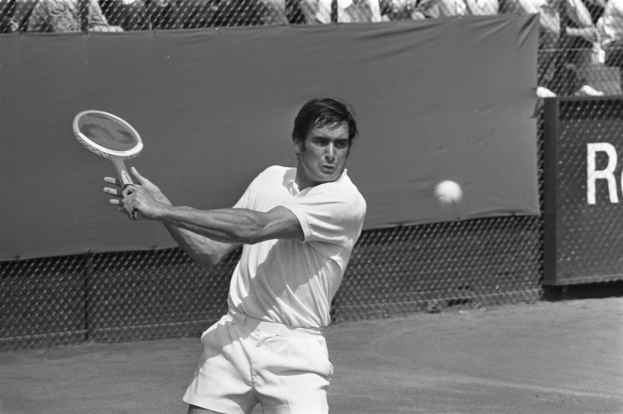 Ein Mann spielt Tennis, schwingt einen Schläger gegen einen Ball, mit Zuschauern im Hintergrund und einem Banner hinter dem Zaun.