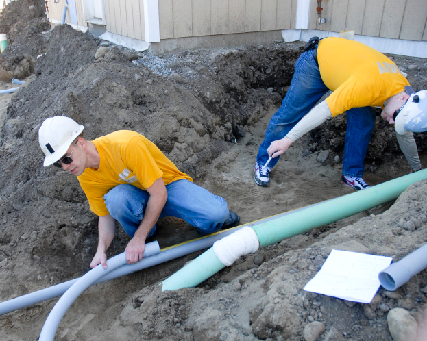 Zwei Männer in gelben Hemden und Helmen arbeiten an einem Rohr, umgeben von Erde, Steinen und anderen Gegenständen, mit einem Gebäude im Hintergrund.