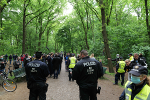 Eine Gruppe von Polizisten steht vor einer Menge von Menschen, einige tragen Mützen und Masken, mit Fahrrädern und einer Bank im Vordergrund und Bäumen und Himmel im Hintergrund, während einer Anti-Terror-Demonstration in Berlin.