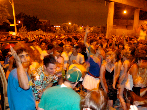Menschen tanzen im Vordergrund bei einem Konzert mit Lichtern, Pfählen, Bäumen und anderen Gegenständen im Hintergrund.