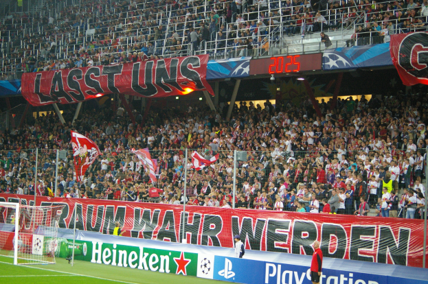 Fußballspiel in einem Stadion mit einer großen Menge, saftig grünem Rasen, einem Torpfosten auf der linken Seite, Bodenbanner, Menschen mit Fahnen und Hintergrundgeländer, Tafeln, Lichter und andere Gegenstände.