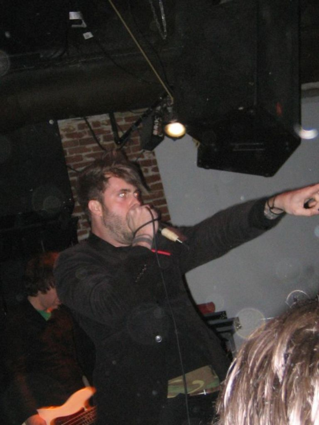 Ein Mann singt in ein Mikrofon in die Mitte einer Gruppe von Menschen, mit Lichtern im Hintergrund.