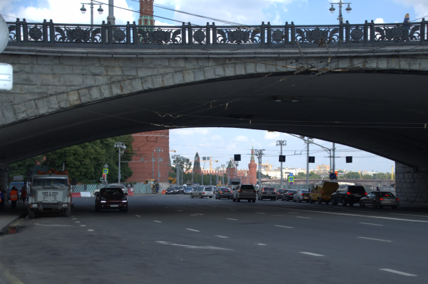 Eine Brücke überspannt eine vielbefahrene Straße mit darunterfahrenden Fahrzeugen, während Fußgänger auf dem Gehweg stehen; Gebäude, Bäume, Laternen, Verkehrssignale und der Himmel sind im Hintergrund sichtbar.