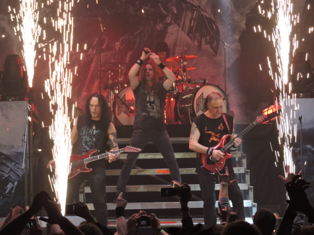 Eine Gruppe von Menschen, die auf der Bühne eines Konzerts spielen, wobei einige Gitarren halten, einer ein Mikrofon und ein anderer Schlagzeug spielt, während das Publikum unten die Performance mit Handys aufzeichnet.