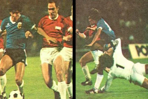 Eine Gruppe von Männern, die auf einem Feld Fußball spielen, wobei einer den Ball tritt und ein anderer versucht, die Kontrolle zu übernehmen, mit einer Tafel im Hintergrund, die auf eine Stadionatmosphäre hindeutet.