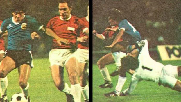 Eine Gruppe von Männern, die auf einem Feld Fußball spielen, wobei einer den Ball tritt und ein anderer versucht, die Kontrolle zu übernehmen, mit einer Tafel im Hintergrund, die auf eine Stadionatmosphäre hindeutet.