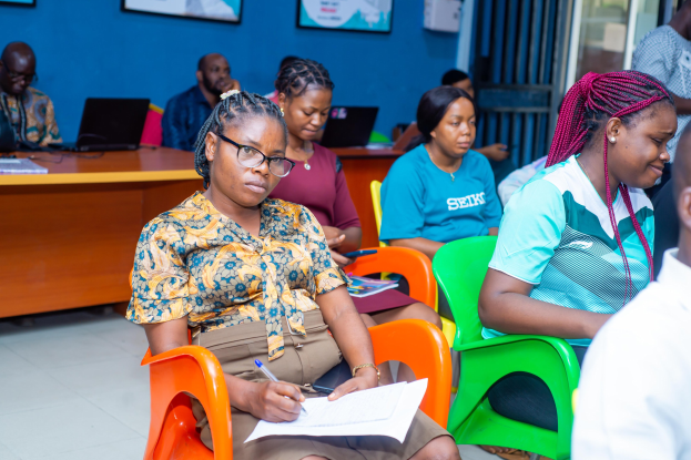 Gruppe von Menschen, die in einem Raum mit Laptops, Büchern und anderen Gegenständen auf Tischen während einer Bildungsveranstaltung in Ghana sitzen, mit gerahmten Gegenständen an der Wand und einer Tür im Hintergrund.