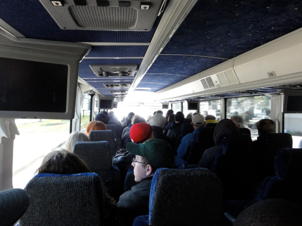 Ein Raum mit vielen sitzenden Menschen, einige tragen Mützen, Fenster mit Vorhängen, Klimaanlagen oben und ein Bildschirm auf der linken Seite.