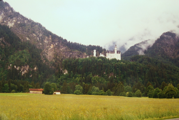 Neuschwanstein Burg in Bayern, Deutschland, umgeben von üppiger Vegetation mit einer Straße, die zu ihr hinaufführt, vor einer Bergkulisse und einem klaren blauen Himmel.