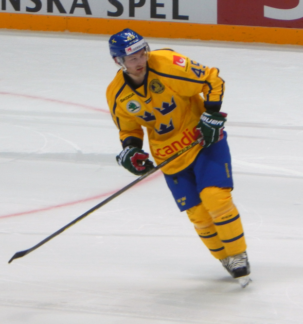 Ein Eishockeyspieler in Bewegung auf dem Eis, der einen Helm, Handschuhe und Schlittschuhe trägt und einen Hockeystock hält, mit einer Tafel im Hintergrund, auf der Text steht.