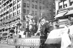 Schwarz-weiß-Foto eines Paradewagens, der mit Statuen von Menschen und Tieren geschmückt ist und an Gebäuden mit Fenstern vorbeifährt, während Zuschauer von der Straße aus zusehen.