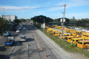 Eine belebte Straße voller Autos, Lastwagen, Bussen, Verkehrskegel, Laternenmasten, Strommasten mit Drähten und Schildern, mit Maschendrahtzaun auf der rechten Seite, Bäumen, Gebäuden und einem bewölkten Himmel im Hintergrund.