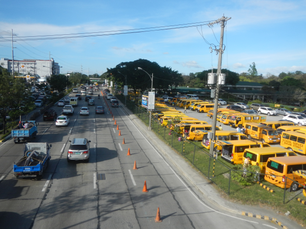 Eine belebte Straße voller Autos, Lastwagen, Bussen, Verkehrskegel, Laternenmasten, Strommasten mit Drähten und Schildern, mit Maschendrahtzaun auf der rechten Seite, Bäumen, Gebäuden und einem bewölkten Himmel im Hintergrund.