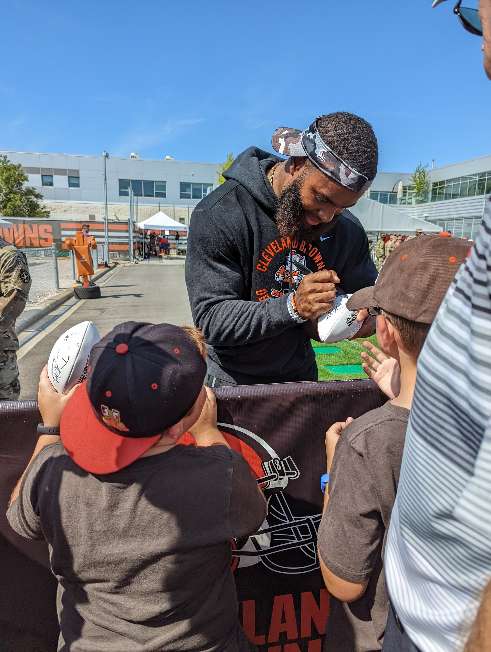 Cleveland Browns Wide Receiver Odell Beckham Jr. signiert Autogramme für Fans auf dem Trainingsgelände, umgeben von Menschen mit Kappen, einem Banner, Gebäuden, Bäumen und Zelten im Hintergrund unter einem bewölkten Himmel.