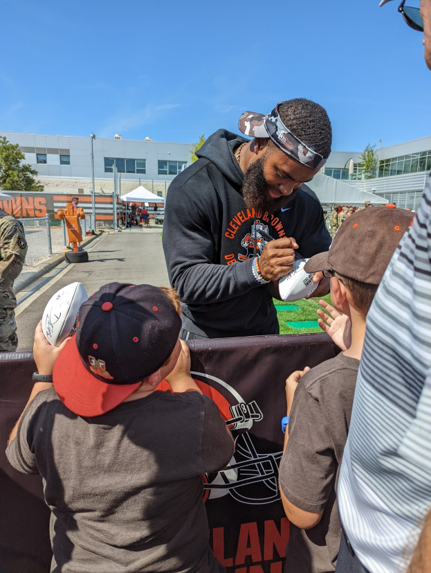 Cleveland Browns Wide Receiver Odell Beckham Jr. signiert Autogramme für Fans auf dem Trainingsgelände, umgeben von Menschen mit Kappen, einem Banner, Gebäuden, Bäumen und Zelten im Hintergrund unter einem bewölkten Himmel.