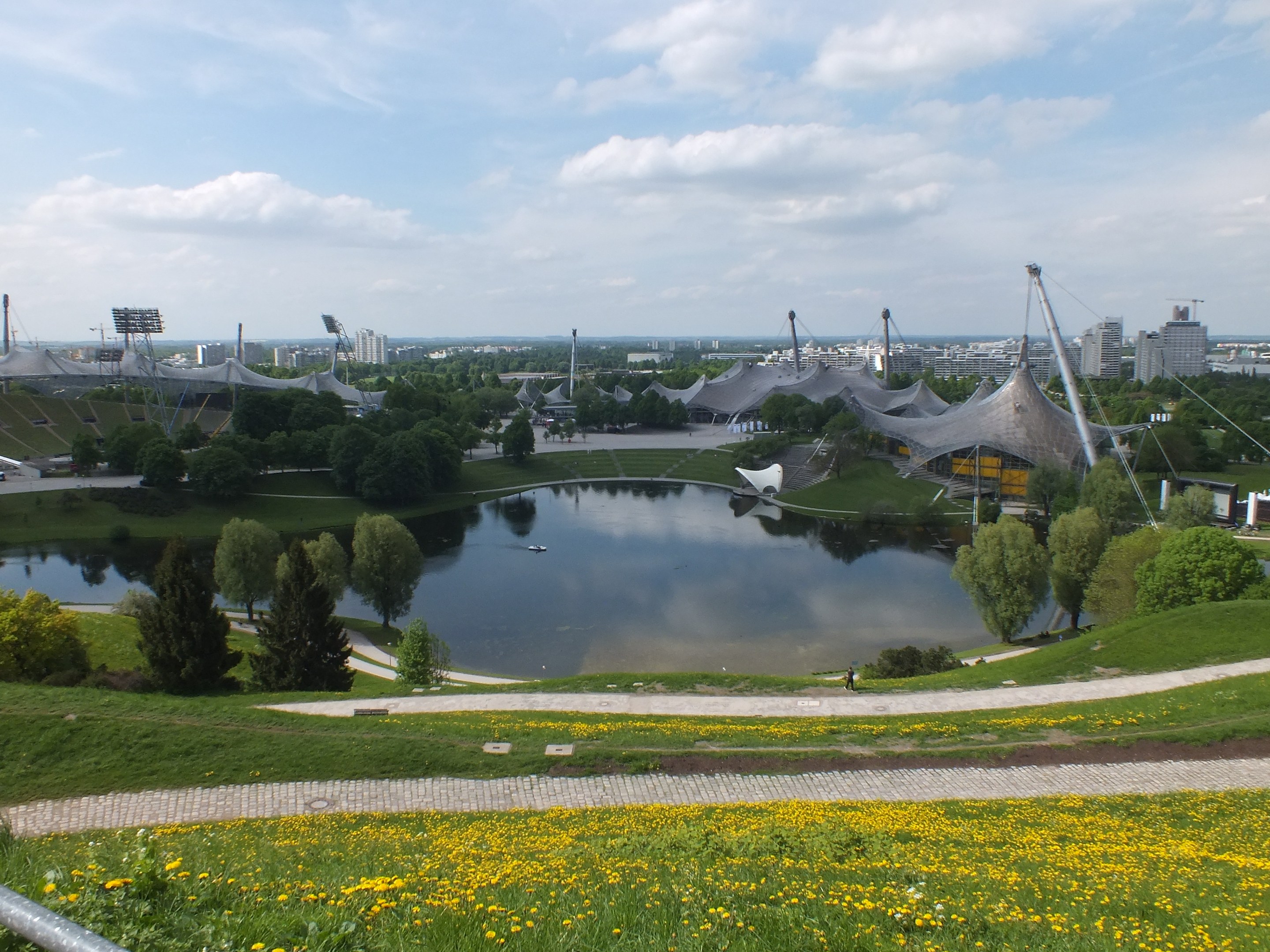 Ansicht des Olympiastadions von einem Hügel aus, mit einem Teich im Vordergrund, der von Grünflächen und gelben Blumen umgeben ist, und Gebäuden im Hintergrund unter einem klaren blauen Himmel.