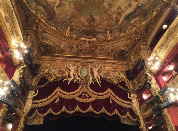 Das Innere eines Theaters mit einem roten Vorhang, Kronleuchtern, Skulpturen, Lichtern und einer Deckenmalerei, das eine Bühne und einige Menschen unten zeigt.