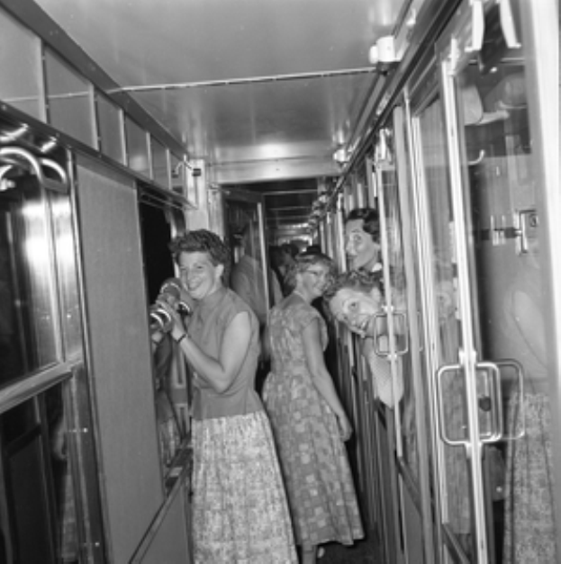 Eine Gruppe von Frauen, die auf einem Zug mit Türen auf beiden Seiten und Lichtern öber ihnen steht, in Schwarz-Weiß dargestellt.