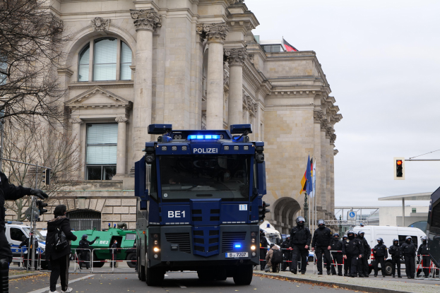 Gruppe von Polizeibeamten vor einem großen Gebäude mit architektonischen Details, Fahrzeugen auf der Straße, einer Person mit einer Kamera, Bäumen, Verkehrsampeln, Fahnenmasten und einem klaren blauen Himmel.