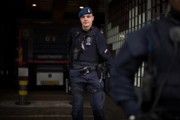 Ein Polizist in Uniform und Mütze steht vor einem Gebäude, rechts daneben eine Person, ein Fahrzeug und Lichter im Hintergrund.