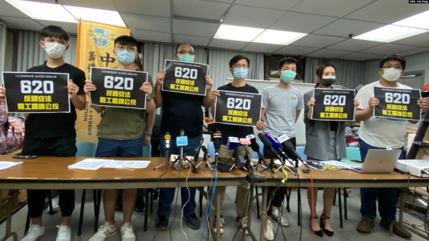 Eine Gruppe maskierter Personen mit Schildern steht vor einem Tisch mit Mikrofonen, Papieren und Laptops und protestiert gegen das Auslieferungsgesetz in Hongkong, mit einem Banner und beleuchteter Decke im Hintergrund.