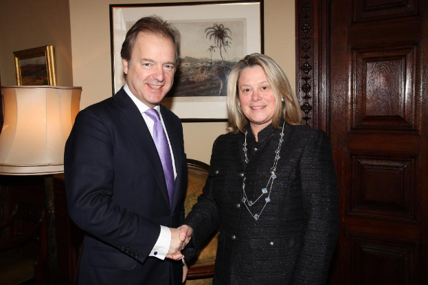 Eine Frau und ein Mann in einem Anzug lächeln, schütteln sich die Hände und sehen sich in einem Raum mit einer Wand, Fotorahmen, einer Lampe, einer Tür und einigen Gegenständen an.