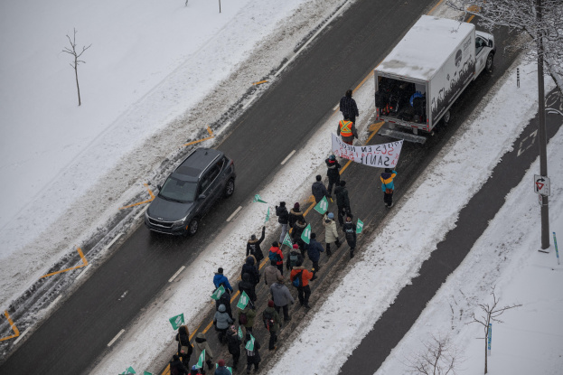 Menschen gehen auf einer verschneiten Straße und tragen Schilder und Plakate, daneben steht ein Lastwagen und ein Auto fährt daneben, Bäume und Pfosten säumen die Straße.