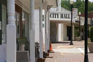 Ein Rathaus umgeben von Gebäuden, Straßenlaternen, Straßenschildern, Autos, Verkehrskegeln, Stühlen, Topfpflanzen, Bäumen und einer bewölkten Himmel