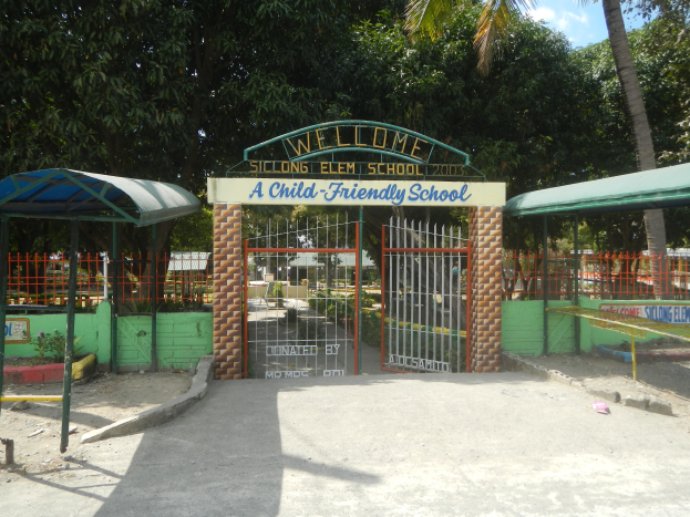 Der Eingang einer kinderfreundlichen Schule mit einem "Willkommen"-Schild oben auf dem Bogen, zwei Unterständen auf beiden Seiten, vielen Bäumen und einem klaren blauen Himmel im Hintergrund.