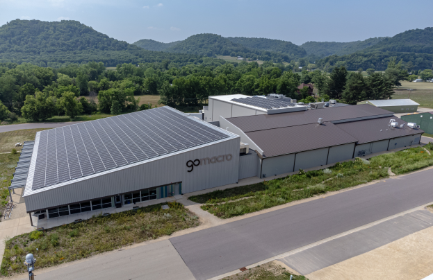Luftaufnahme des Go Macro Solar Power Plant, ein großes Gebäude mit Solarmodulen auf dem Dach, umgeben von Grünflächen und einer Straße, mit Hügeln und einem klaren blauen Himmel im Hintergrund.