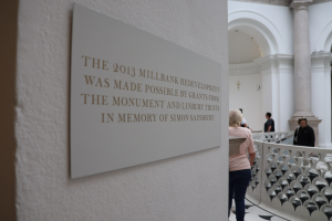 Gruppe von Menschen in einem Innenraum mit einer Wandtafel, einem Zaun, Säulen, Deckenleuchten und einem Schild über die 2013er Millbank-Renovierung, die von den Monument- und Linbury Trusts sowie in Erinnerung an Simon Sainsbury finanziert wurde.