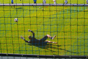 Ein Fussballer hält den Ball vor einem Tor, mit einem Netz im Vordergrund und einigen Menschen im Hintergrund sowie mehreren Bällen auf dem Boden verstreut.