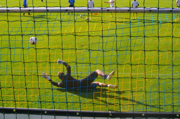 Ein Fussballer hält den Ball vor einem Tor, mit einem Netz im Vordergrund und einigen Menschen im Hintergrund sowie mehreren Bällen auf dem Boden verstreut.
