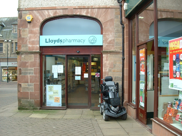 Eine Apotheken-Filiale mit einem vor ihr geparkten Fahrzeug und einem im linken Winkel sichtbaren Geb├Ąude.