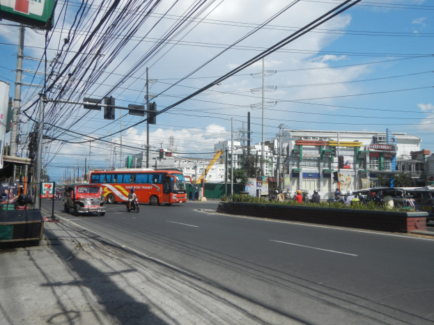 Eine belebte Stadtstraße mit Fahrzeugen, Strommasten, Ampeln, Schildern, Bäumen, Gebäuden und einem bewölkten Himmel.